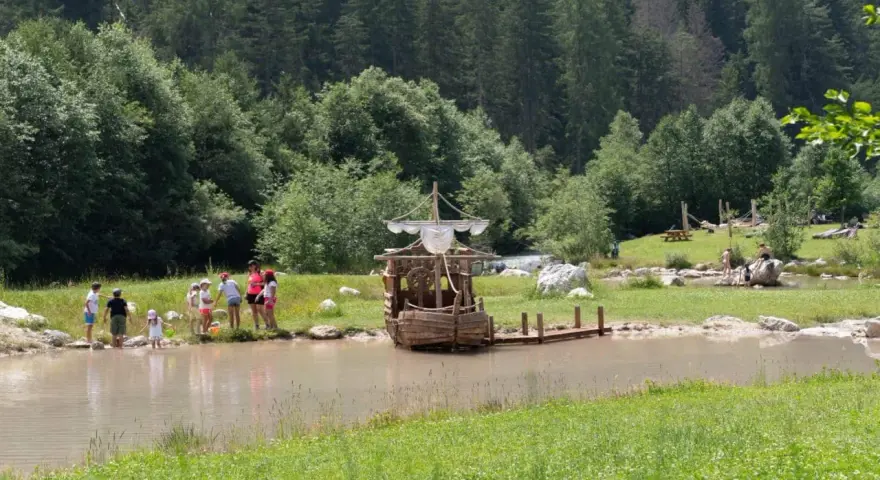 Bambini giocano vicino a un galeone di legno in un lago, mentre altri si rilassano sull'erba. Perfetto per le **san vigilio di marebbe passeggiate** estive.
