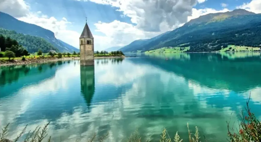 Torre sommersa nel lago di Resia, con montagne verdi e cielo nuvoloso.