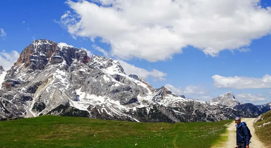 Un uomo con bastoncini da trekking su un sentiero in Trentino, con imponenti montagne innevate sullo sfondo. Un'esperienza di trekking trentino facile e panoramica.
