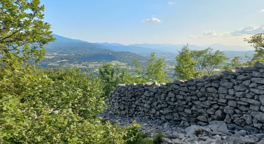 Muro di pietra a secco con vista panoramica su una valle alpina e montagne all'orizzonte.