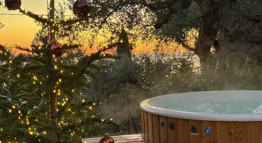 Un albero di Natale illuminato e una vasca idromassaggio fumante al tramonto sul Lago di Garda. Il camping è aperto tutto l'anno, un'oasi di relax invernale.