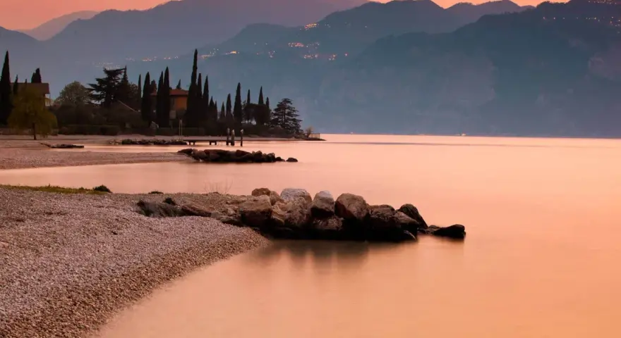 Brenzone sul Garda: cosa vedere? Spiaggia di ciottoli, cipressi e montagne al tramonto sul lago.