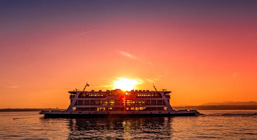 Traghetto in navigazione sul Lago di Garda al tramonto, con il sole che illumina il cielo e il riflesso sull'acqua.