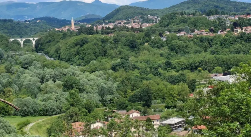 Panoramica collinare con un ponte ad archi, un campanile e case sparse a Cassano Magnago, circondata da rigogliosa vegetazione.