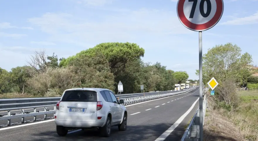 Un'auto bianca percorre una strada extraurbana con limite di 70 km/h. Alberi e cielo azzurro fanno da cornice.