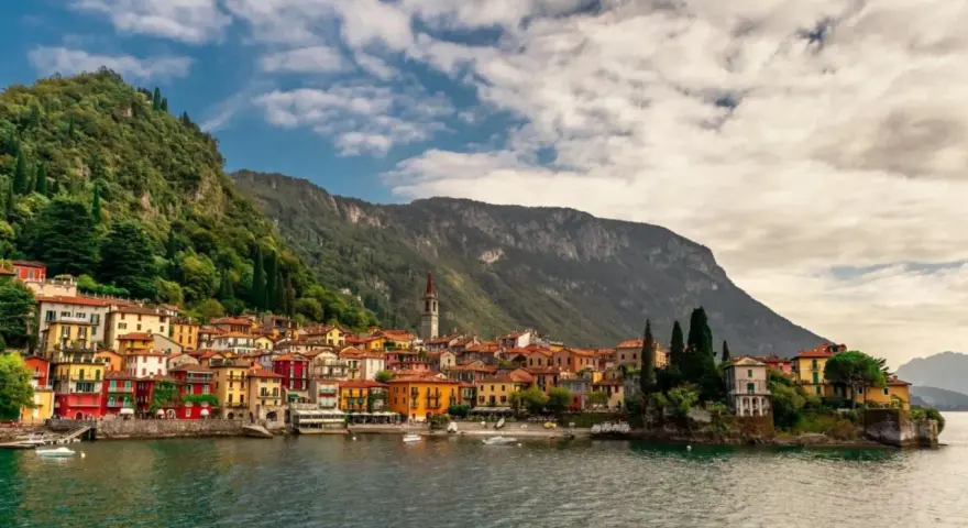 Colorate case affacciate sul lago di Lecco, con montagne verdi sullo sfondo e un cielo nuvoloso.
