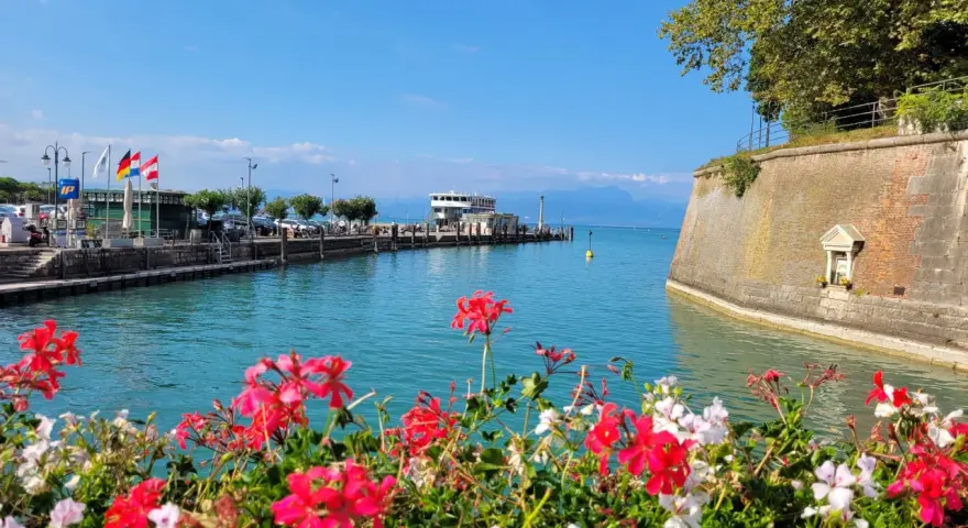 Fiori rossi e bianchi in primo piano, con un molo, un traghetto e montagne sullo sfondo. Un'immagine che evoca i paesi vicino Peschiera del Garda.