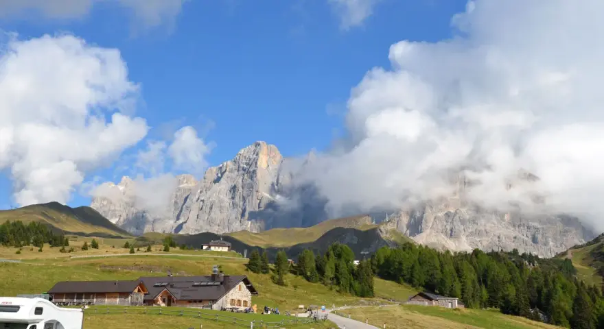 Panorami mozzafiato al Passo Rolle: malghe, boschi e vette imponenti sotto un cielo azzurro con nuvole bianche.