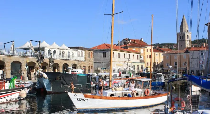 Porto di Muggia cosa vedere: barche a vela, edifici storici e un campanile svettano sotto il cielo azzurro.