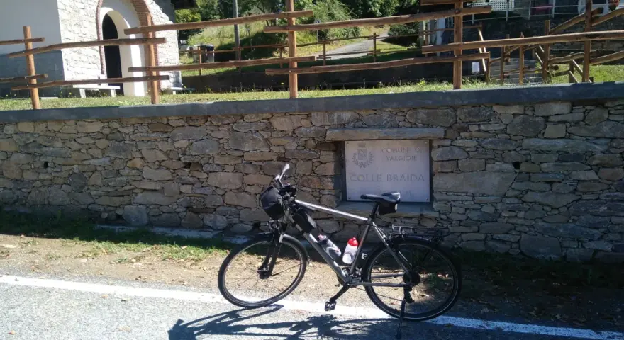 Bicicletta ferma sul ciglio della strada, con un muretto in pietra e un cartello che indica "Colle Braida".