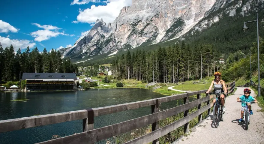 Famiglia in bicicletta sul ponte del **lago di San Vito**, con montagne imponenti e boschi sullo sfondo.