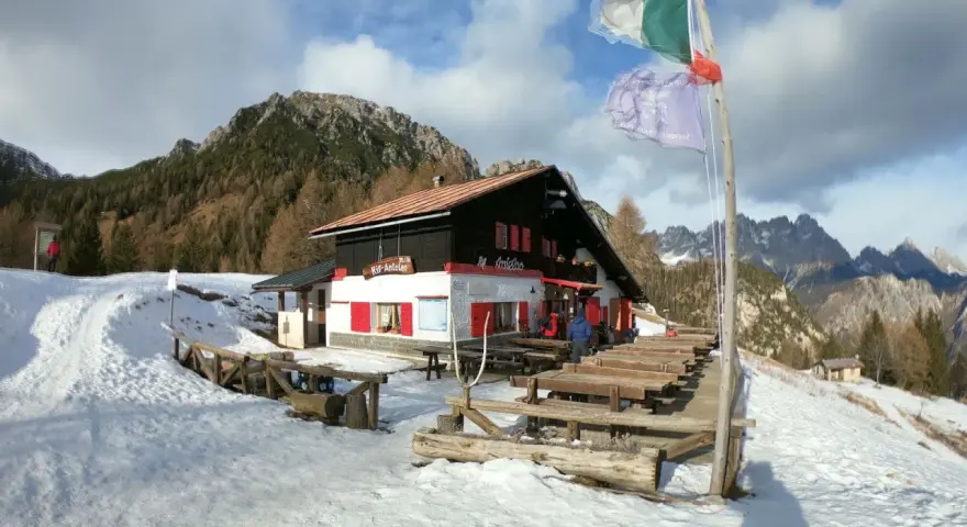 Rifugio Antelao innevato, con tavoli esterni e bandiere. Informazioni su come arrivare al rifugio sono visibili.