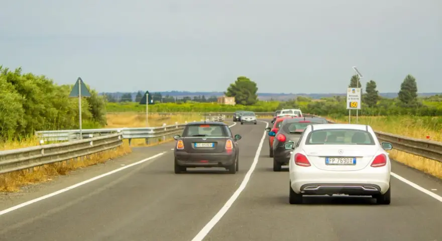 Traffico in colonna su strada con vegetazione ai lati. Un'auto tenta il sorpasso linea continua, mettendo a rischio la sicurezza.