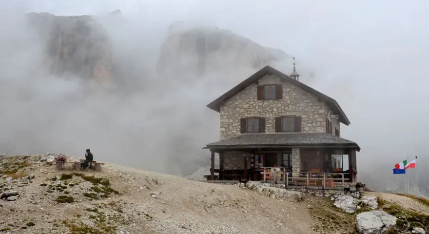 Rifugio Franz Kostner avvolto dalla nebbia, con un escursionista solitario su una panchina e le bandiere che sventolano.