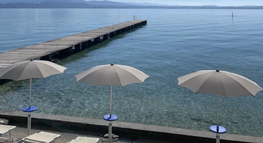 Ombrelloni e lettini al lido delle Bionde, con un pontile in legno che si estende sul lago limpido e montagne all'orizzonte.