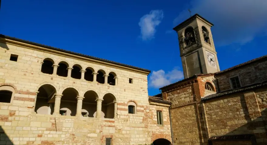 Antica **cella monte** con portici ad archi e torre campanaria sotto un cielo azzurro.