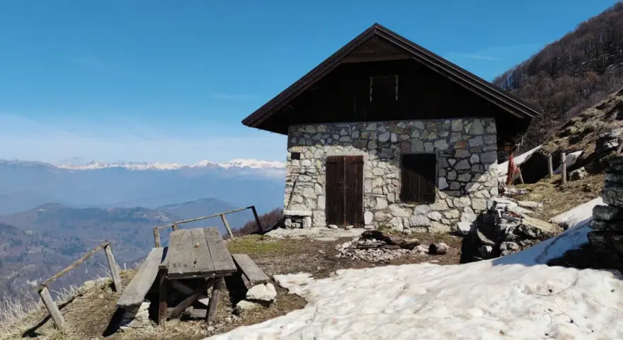 Bivacco Monte Grappa, rifugio in pietra con tavolo da picnic e vista sulle montagne innevate.