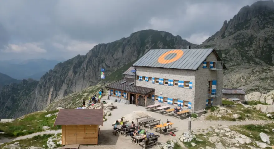 Il rifugio Cima d'Asta, con le sue finestre blu e arancioni, accoglie escursionisti tra rocce e montagne nebbiose.