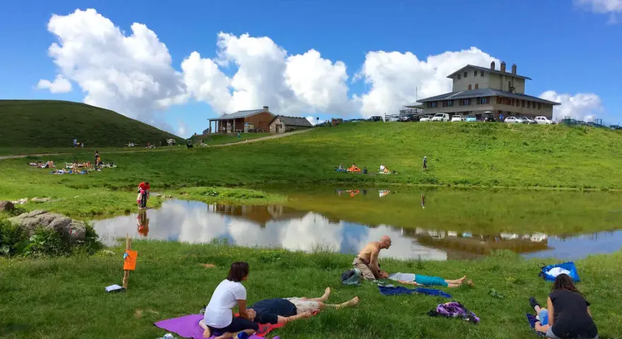 Persone rilassate sui prati verdi, vicino a un lago, sotto un cielo azzurro con nuvole bianche. Sembra un luogo ideale per i piani dell'avaro, godendosi la natura.