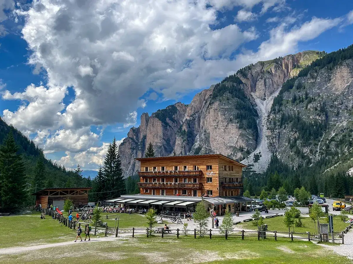 Un hotel in legno ai piedi di imponenti montagne, con un prato verde e persone che passeggiano. Vicino al lago verde San Vigilio di Marebbe.