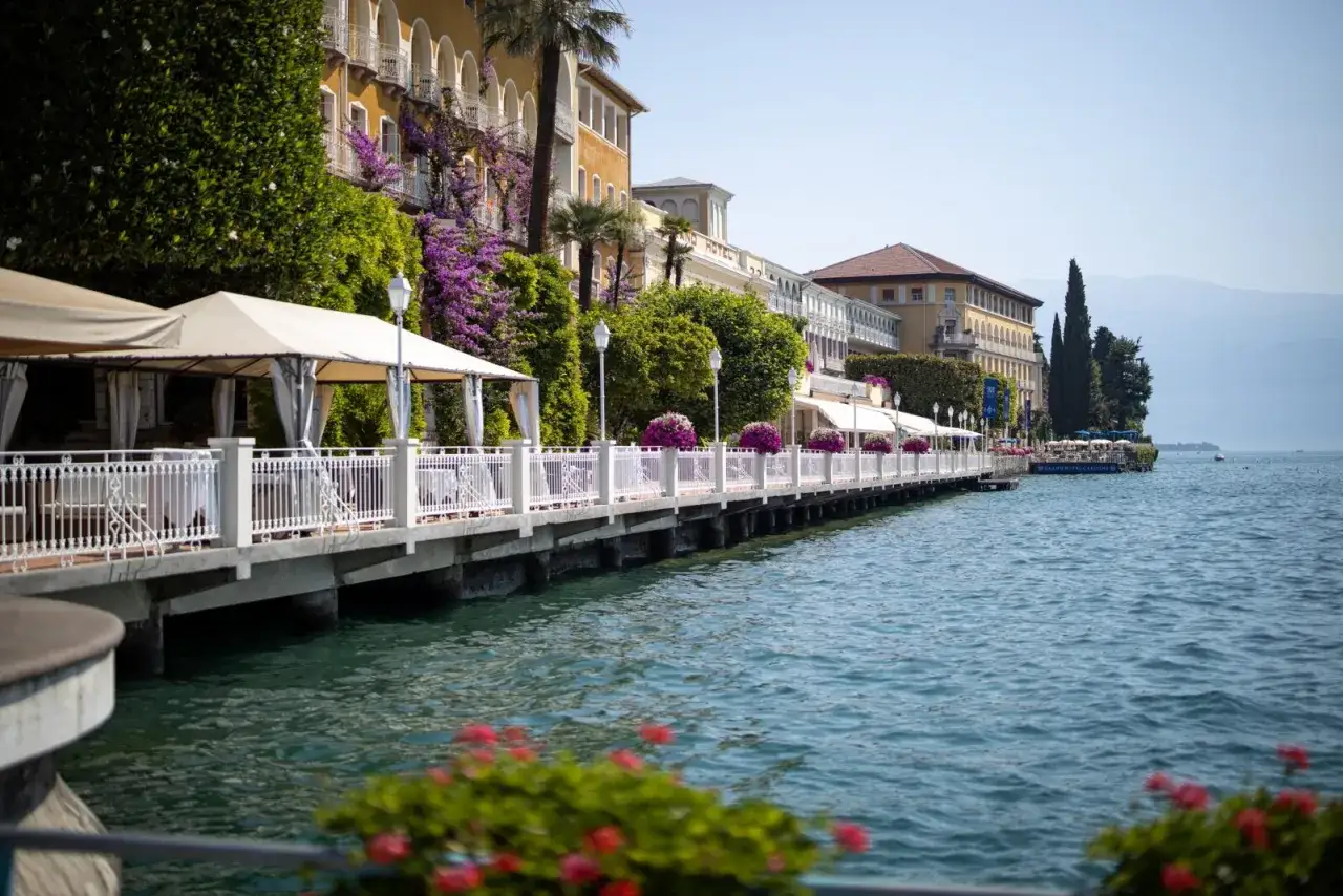 Passeggiata sul lago a Gardone Riviera centro storico, con hotel storici, giardini fioriti e tavoli all'aperto.