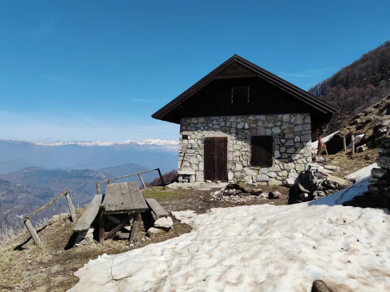 Bivacco Monte Grappa, un rifugio in pietra con tavolo da picnic e vista sulle montagne innevate.