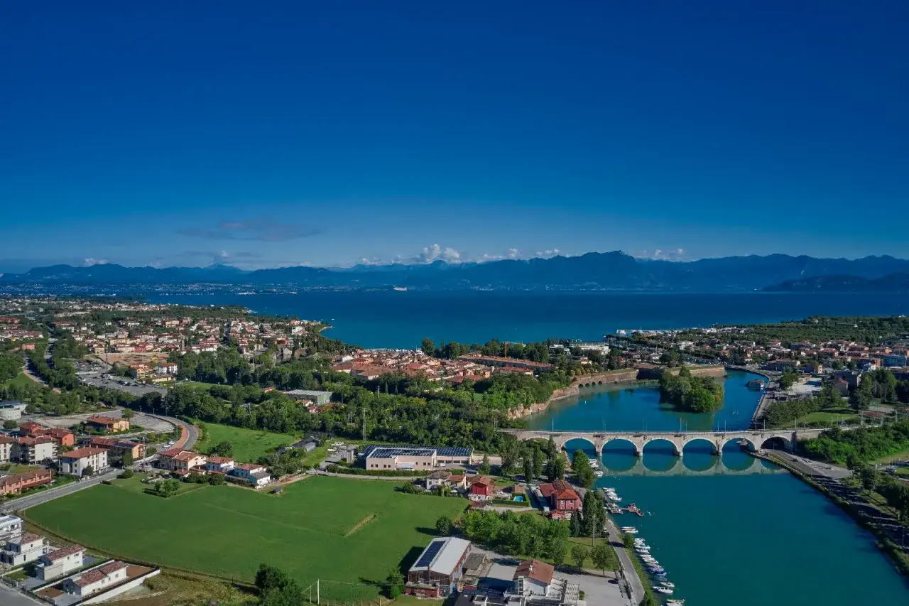 Vista aerea di Peschiera del Garda, dove il fiume Mincio incontra il Lago di Garda, con ponti, case e montagne sullo sfondo.
