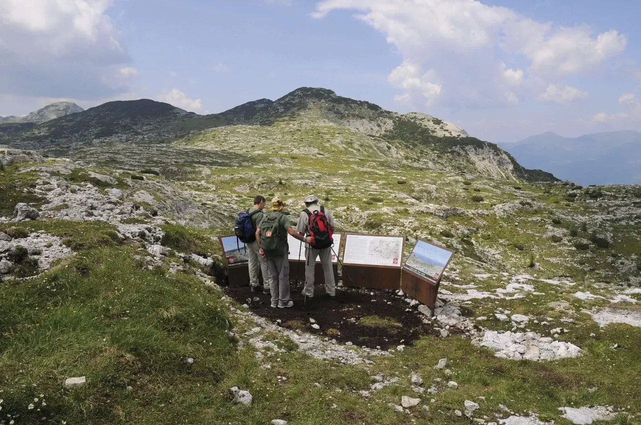 Escursionisti leggono pannelli informativi sul monte Ortigara come arrivare. Paesaggio montano roccioso e verdeggiante.