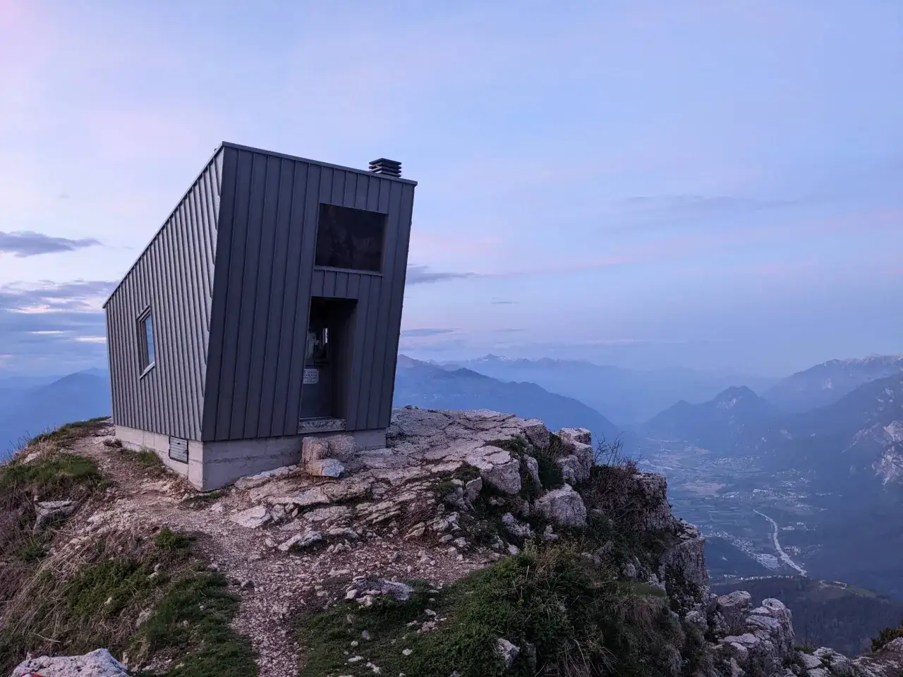 Bivacco Giacomelli alla Madonnina, un rifugio moderno su una cresta rocciosa con vista panoramica su una valle alpina al tramonto.
