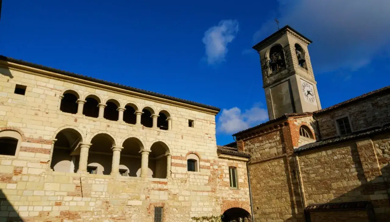 Antica cella monte con portici ad archi e campanile con orologio, sotto un cielo azzurro intenso.