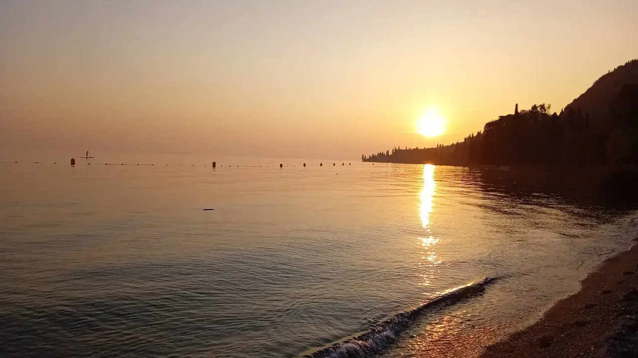 Tramonto dorato sul Lago di Garda, un weekend perfetto con un SUPista solitario e il riflesso del sole sull'acqua calma.