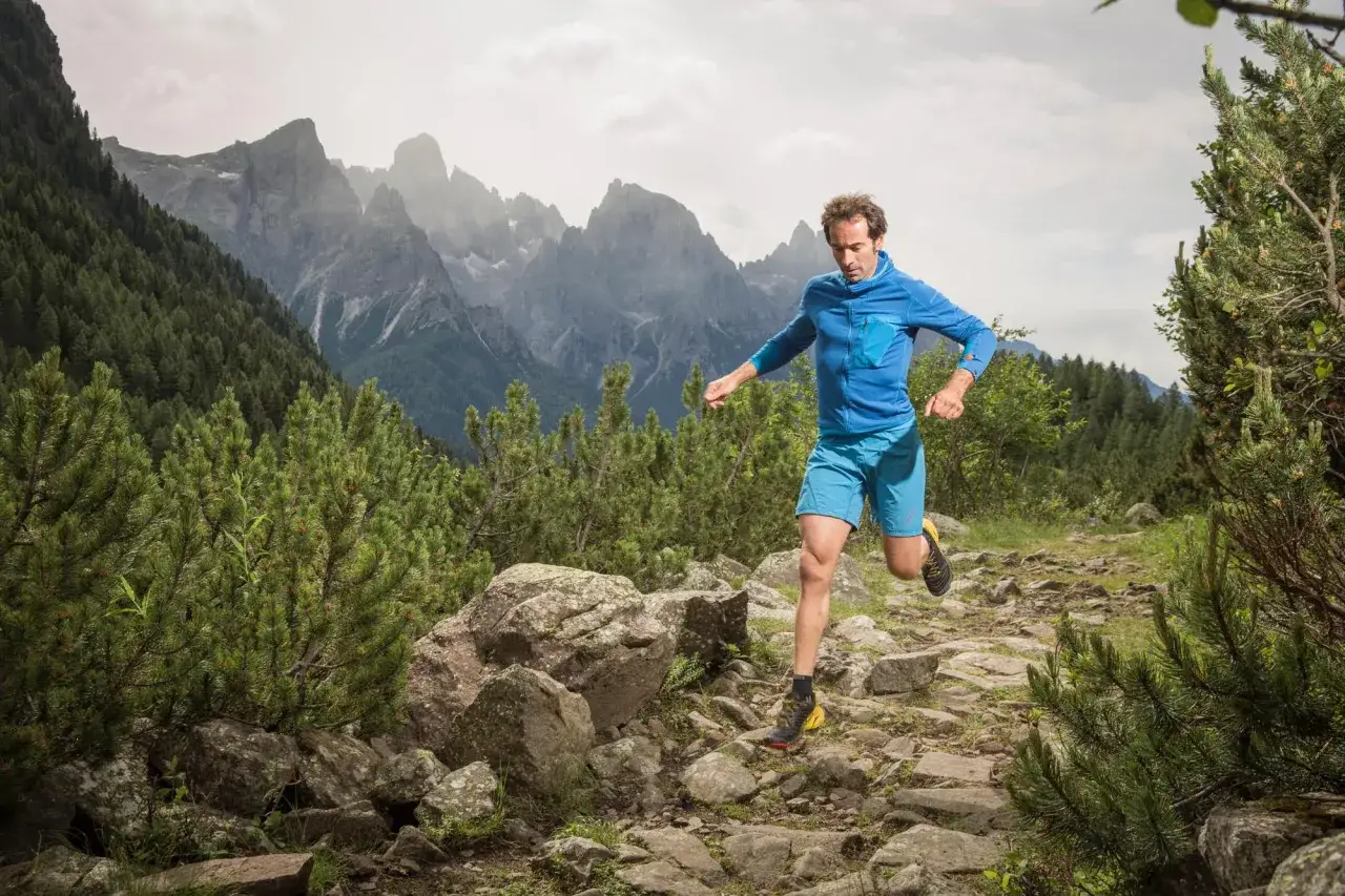 Un trail runner in blu corre su un sentiero roccioso tra pini e montagne maestose. Per raggiungere malga Valcigolera, segui questo percorso panoramico.