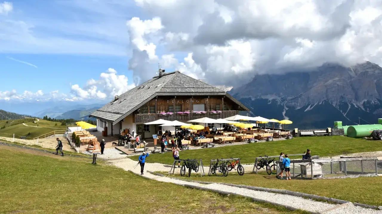 Rifugio Pralongi&agrave;, con tavoli all'aperto e montagne sullo sfondo. Per sapere come arrivare, guarda i sentieri e le bici.