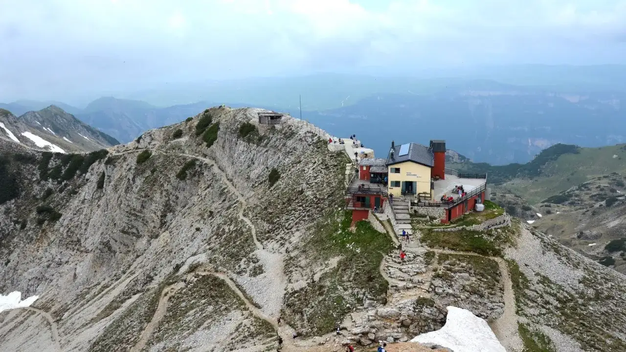 Sentiero per il rifugio Fraccaroli, un edificio giallo su una cresta rocciosa con sentieri tortuosi e persone che camminano.
