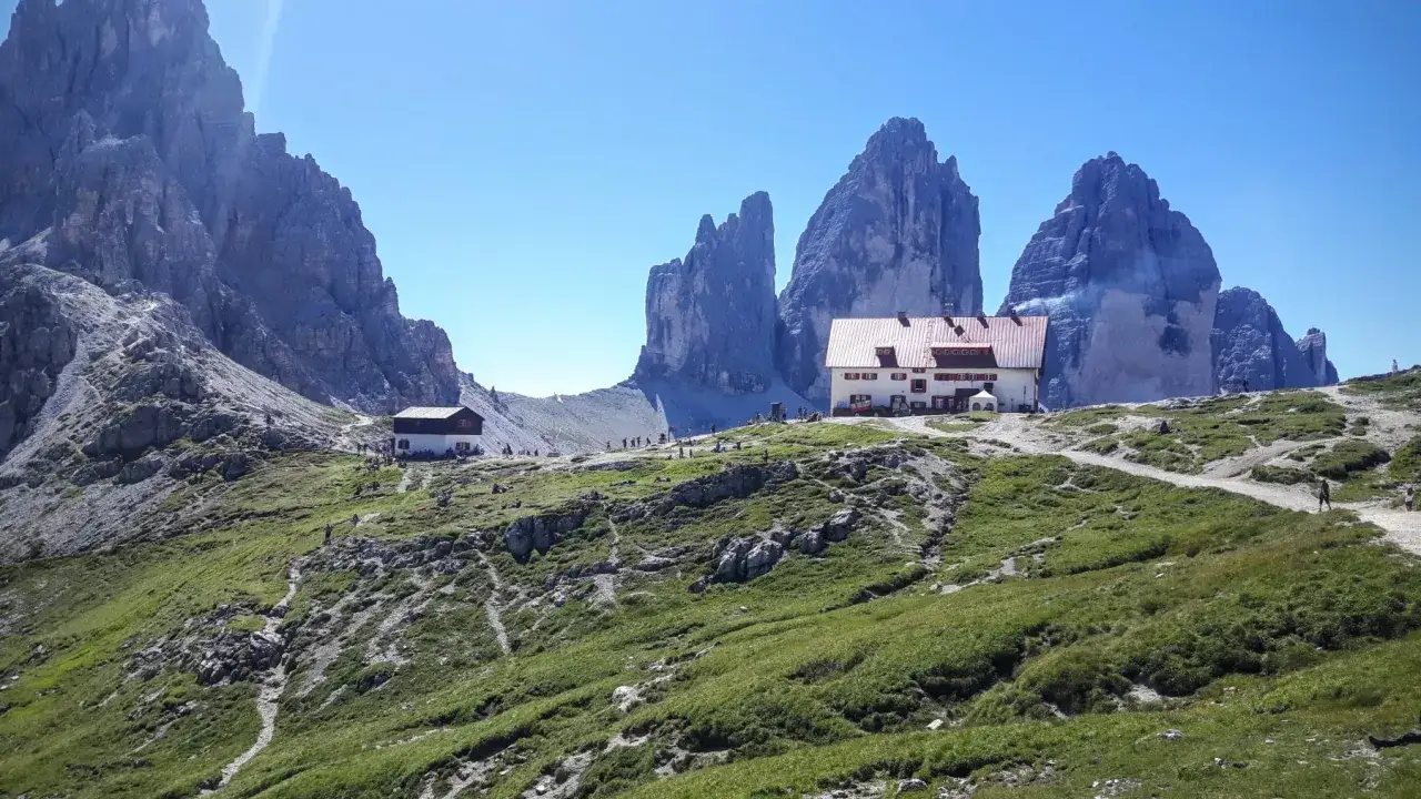 Il rifugio Locatelli, con le Tre Cime di Lavaredo sullo sfondo, è un punto di ristoro per escursionisti in un paesaggio alpino verdeggiante.