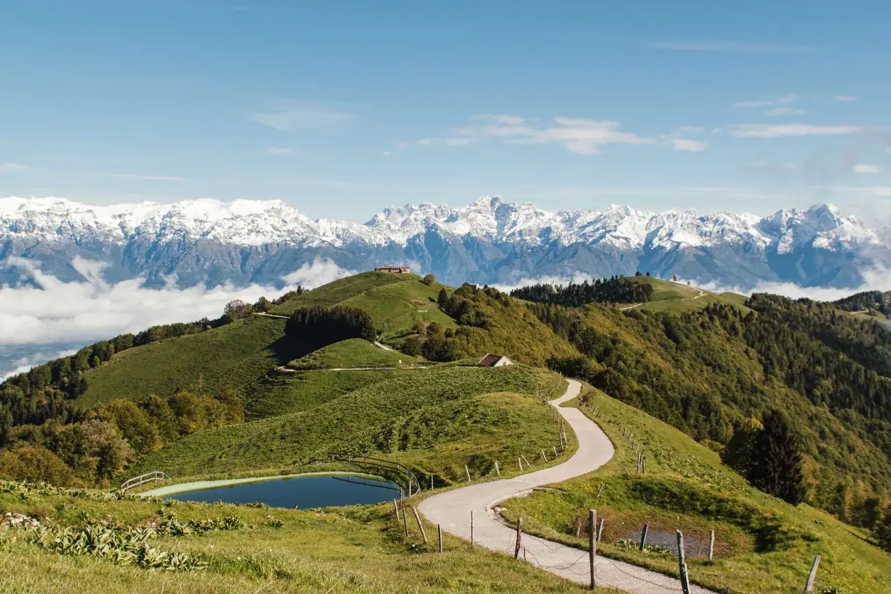 Malga Gard&agrave; Lentiai: sentiero serpeggiante tra verdi colline, laghetto alpino e maestose cime innevate all'orizzonte.