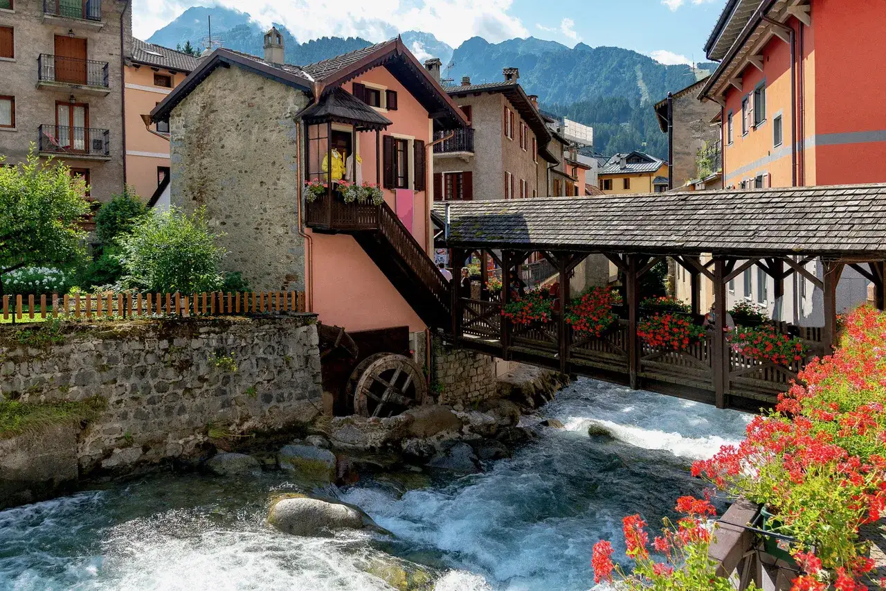 Un pittoresco ponte di legno coperto attraversa un torrente impetuoso, fiancheggiato da case colorate e fiori rossi. Un mulino ad acqua antico aggiunge fascino a questo luogo da cartolina, perfetto per scoprire cosa vedere.