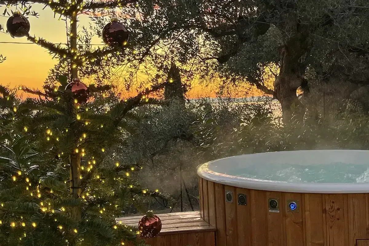 Un albero di Natale illuminato e palline rosse vicino a una vasca idromassaggio fumante al tramonto sul Lago di Garda. Il camping &egrave; aperto tutto l'anno.