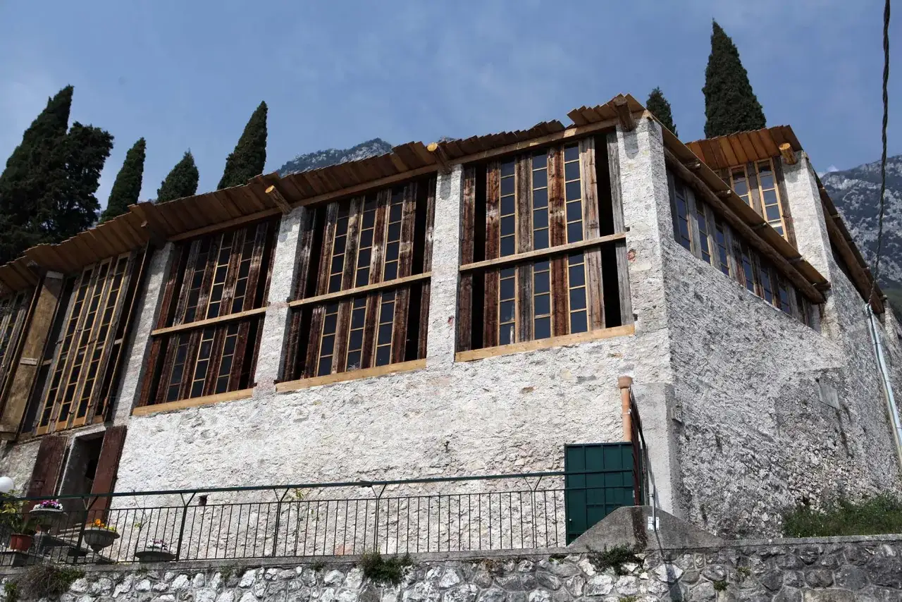 Edificio in pietra con ampie finestre in legno, circondato da cipressi e montagne. Un luogo che evoca la bellezza dei limoni sul Garda.