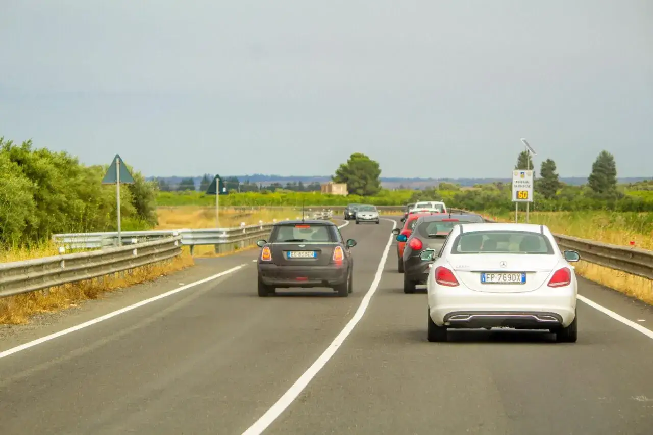 Traffico in coda, un'auto tenta un sorpasso linea continua. Auto in colonna su strada con vegetazione ai lati.