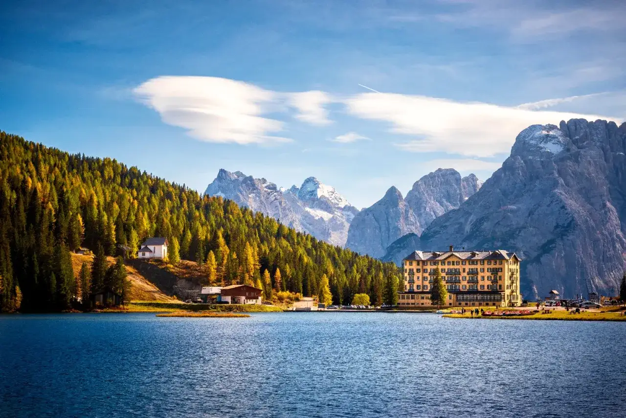 Splendido lago alpino tra Veneto e Trentino, con boschi autunnali, montagne innevate e un grande albergo giallo.