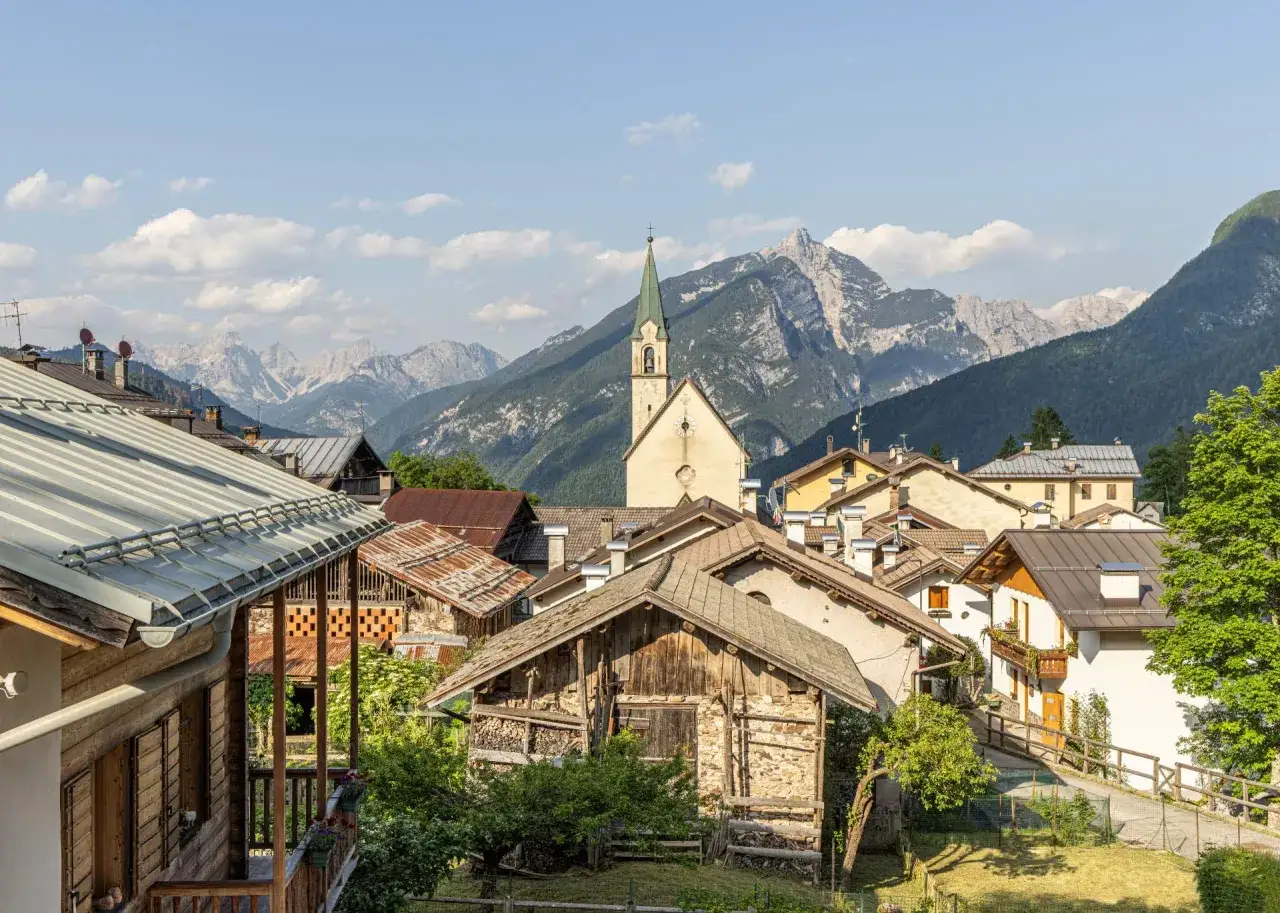 Sagron Mis: un borgo alpino con case in pietra e legno, una chiesa con campanile e maestose montagne sullo sfondo.