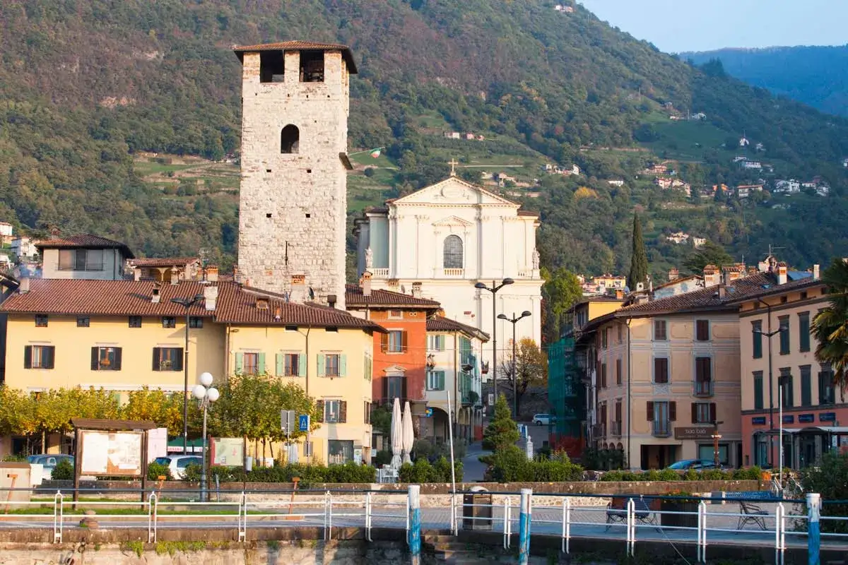 Pisogne: torre medievale, chiesa bianca e case colorate affacciate sul lago, con montagne verdi sullo sfondo. Cosa vedere in questo borgo incantevole.