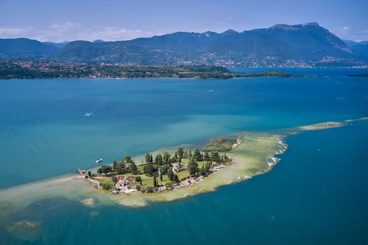 L'Isola dei Conigli sul Lago di Garda, un gioiello verdeggiante con case e moli, circondata da acque turchesi e montagne sullo sfondo.