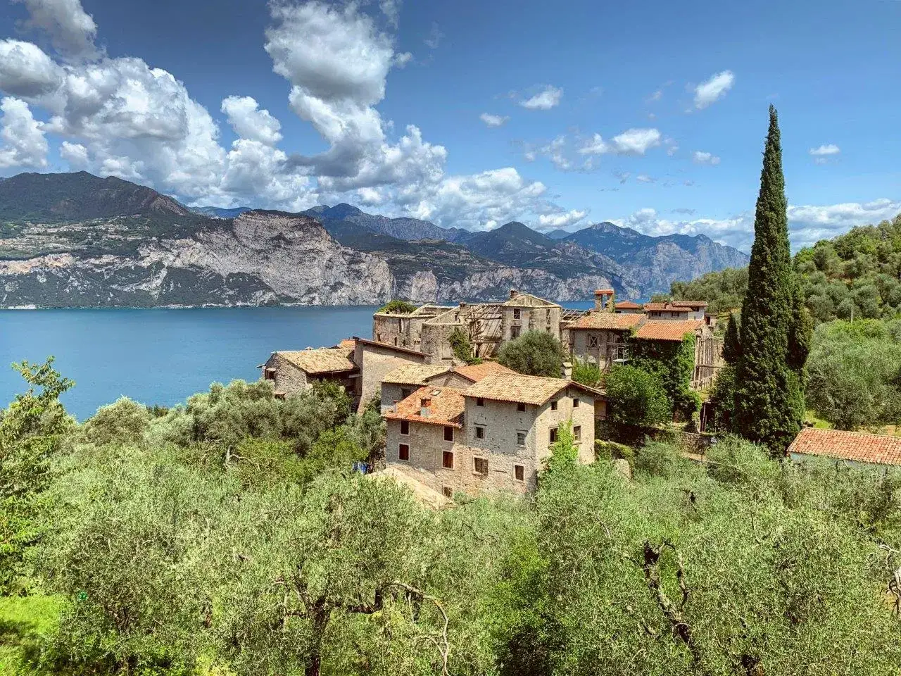 Borgo di Brenzone sul Garda, con case in pietra e ulivi secolari, si affaccia sul lago azzurro e le montagne.