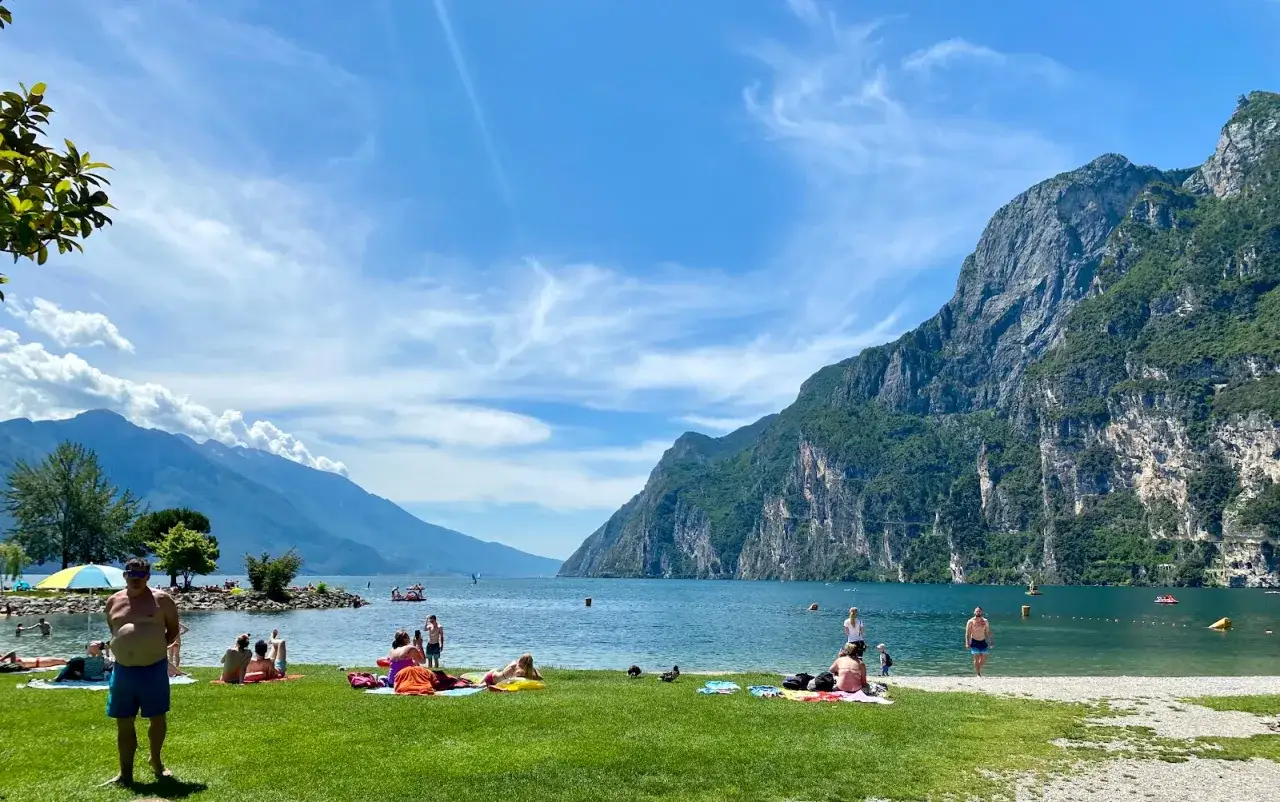 Giornata di sole sulla spiaggia sabbioni riva del garda. Gente rilassata sull'erba e in acqua, montagne imponenti sullo sfondo.