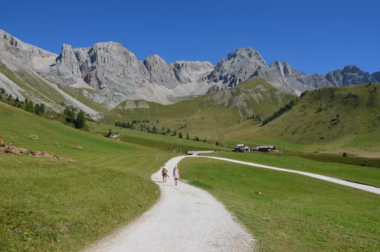 Sentiero panoramico per canazei escursioni facili, con due persone che camminano verso rifugi alpini e maestose montagne sullo sfondo.
