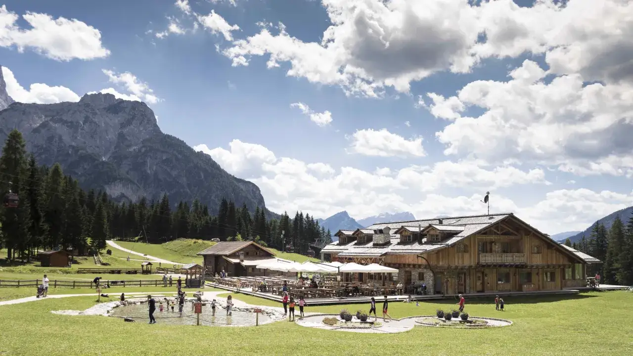 Un rifugio alpino con tavoli all'aperto e un laghetto dove giocano bambini, incorniciato da montagne maestose. Un'oasi di pace ai Piani di Pezz&egrave;.