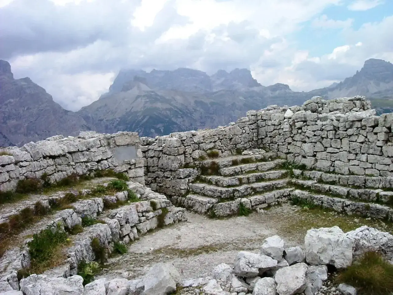 Rovine in pietra sul Monte Piana, con imponenti montagne sullo sfondo e nuvole minacciose.
