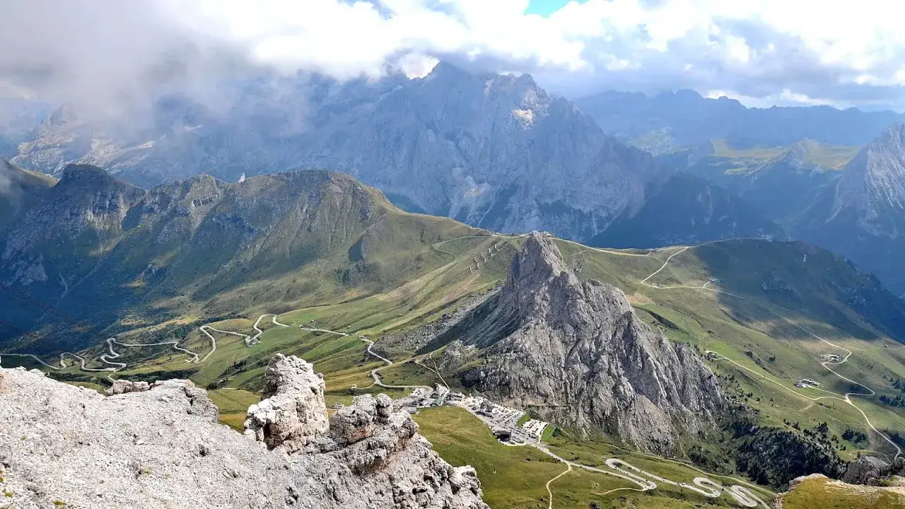 Panoramica mozzafiato del Passo Pordoi con strade tortuose che si snodano tra verdi pendii e imponenti vette rocciose, sotto un cielo nuvoloso.